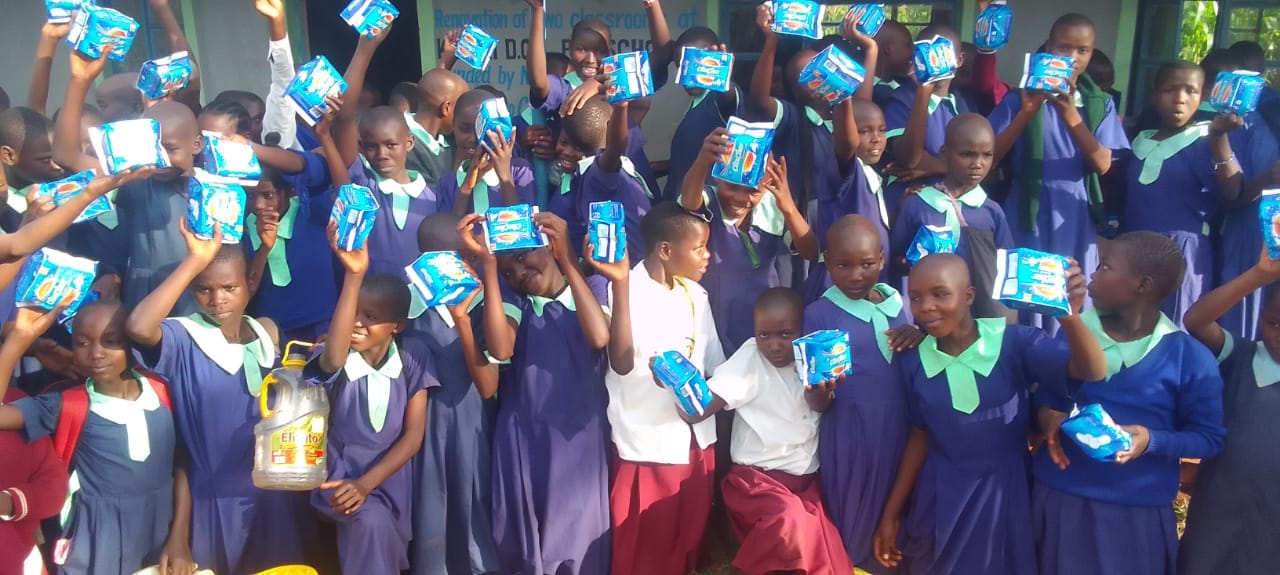 A close group of pupils in purple school pinafores holding packets of sanitary pads above their heads.