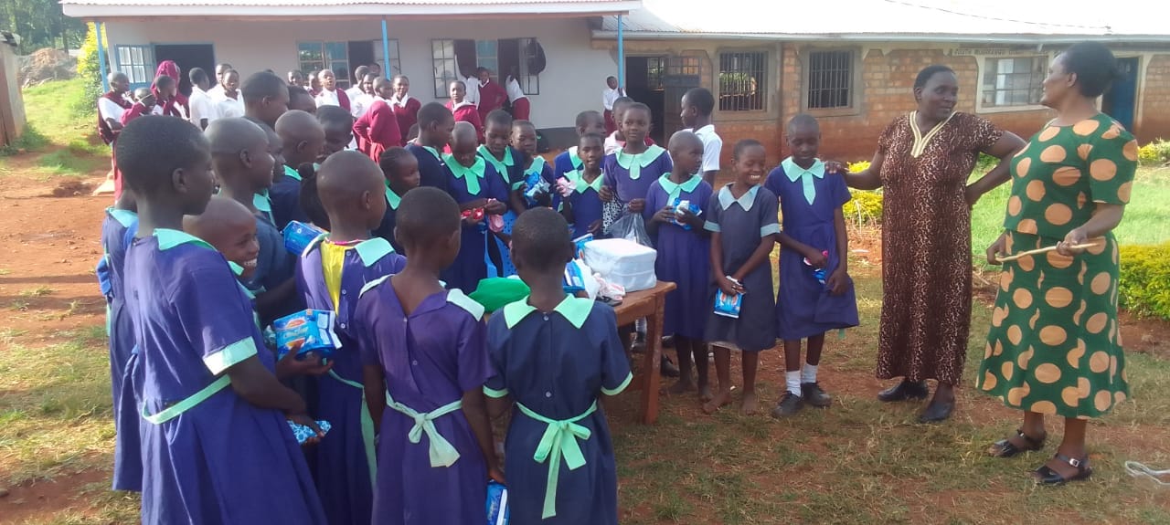 Pupils queuing at a table outside a classroom to collect packets of sanitary pads, with two women from the organising committee standing alongside.