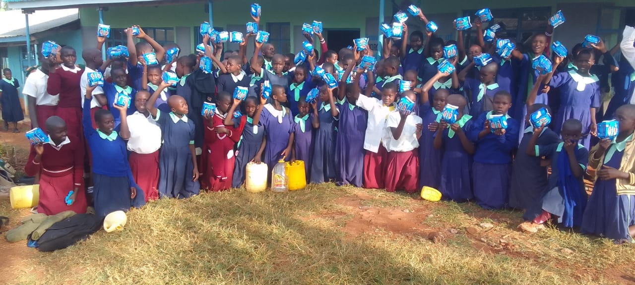 A large group of pupils outside their school building raising packets of sanitary pads.