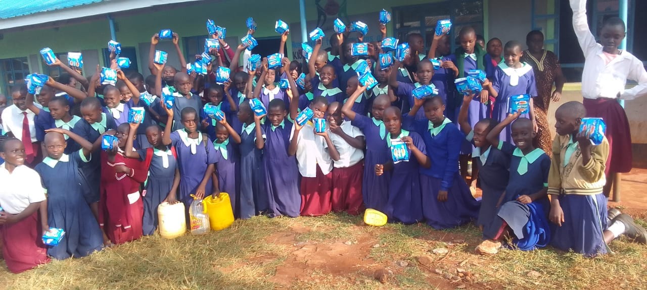 Pupils outside the school building celebrating with packets of sanitary pads raised above their heads.
